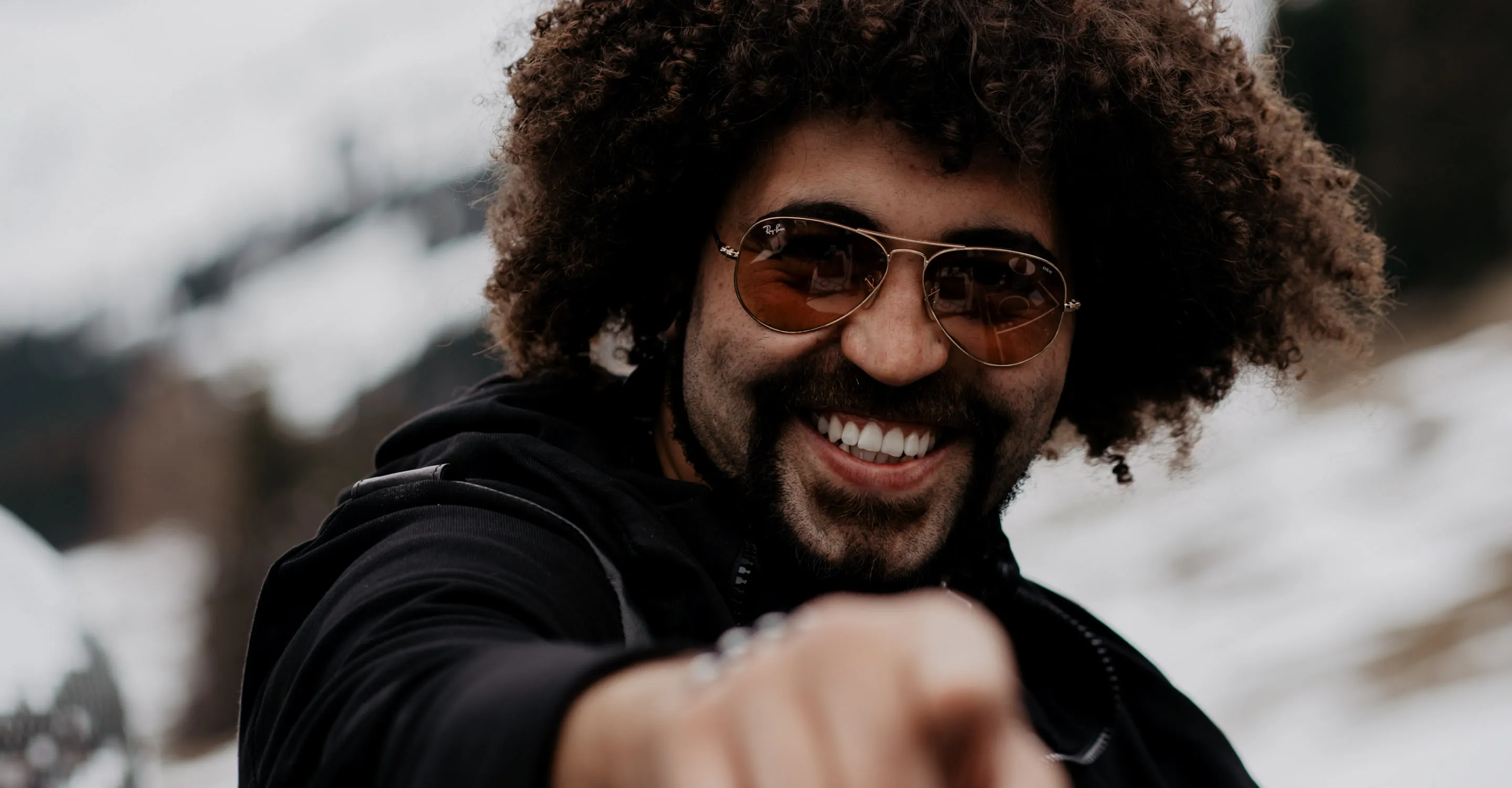 Person with curly hair and sunglasses smiling and pointing outdoors