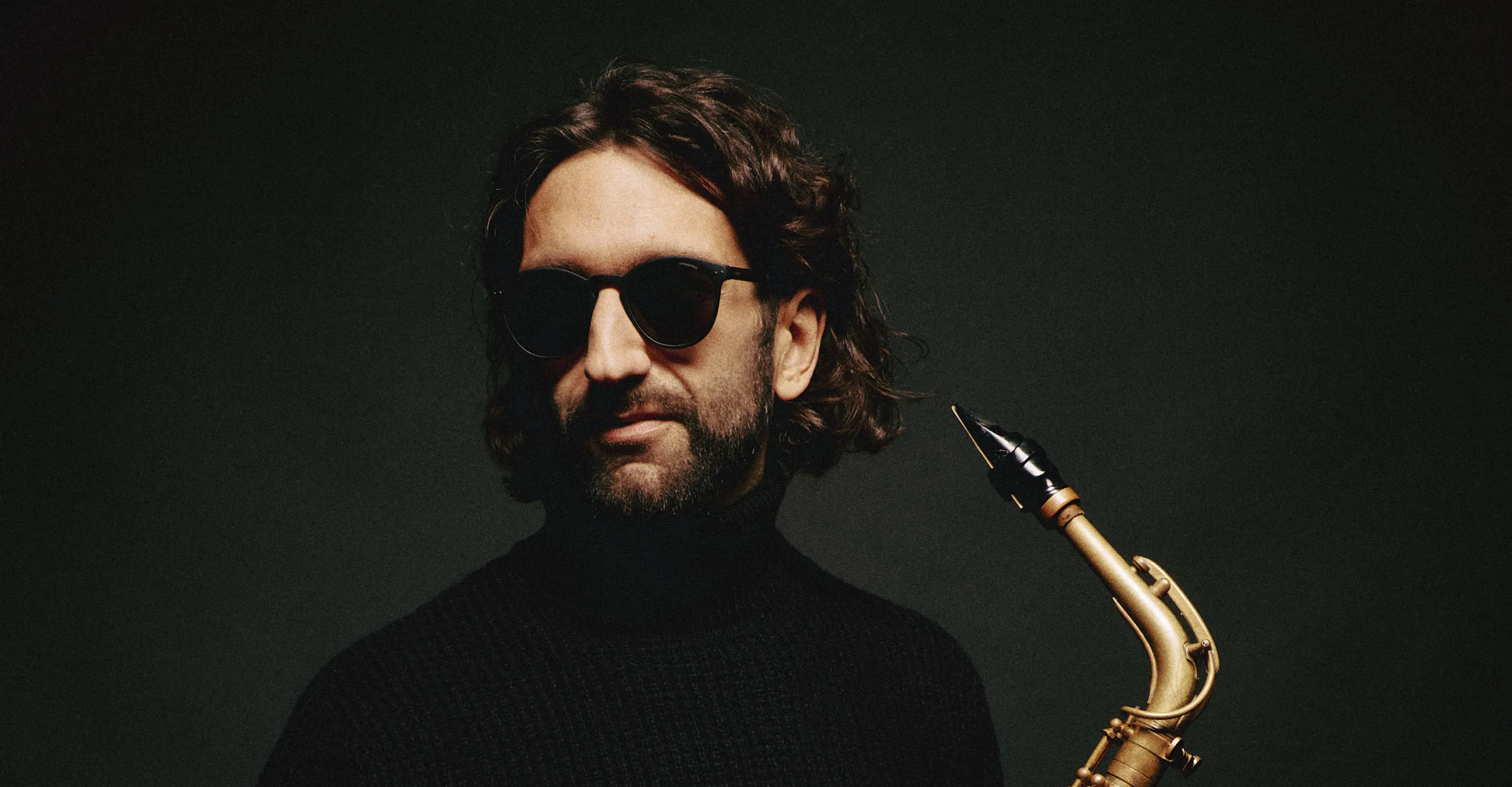 Man with sunglasses holding a saxophone against a dark background