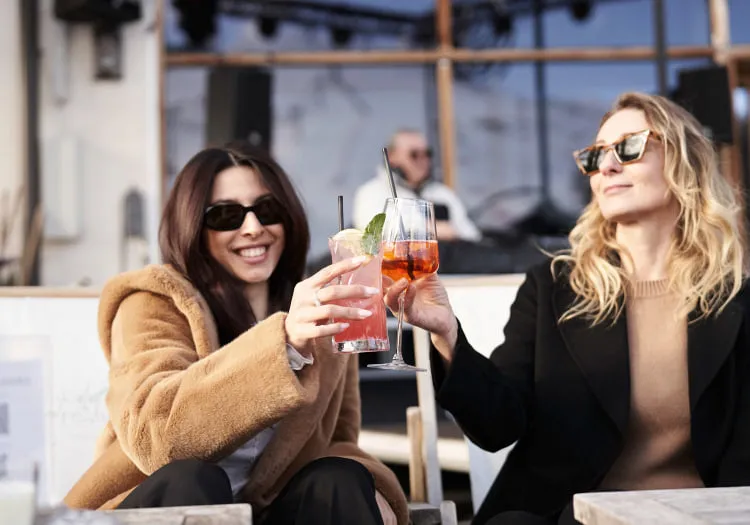 Zwei Frauen heben Gläser in einem Holzhotel., im Hintergrund ein DJ.