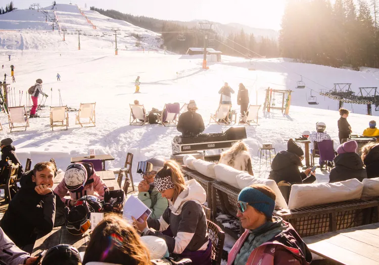 Menschen auf einer Terrasse in den Bergen, Schnee und Skifahrer im Hintergrund