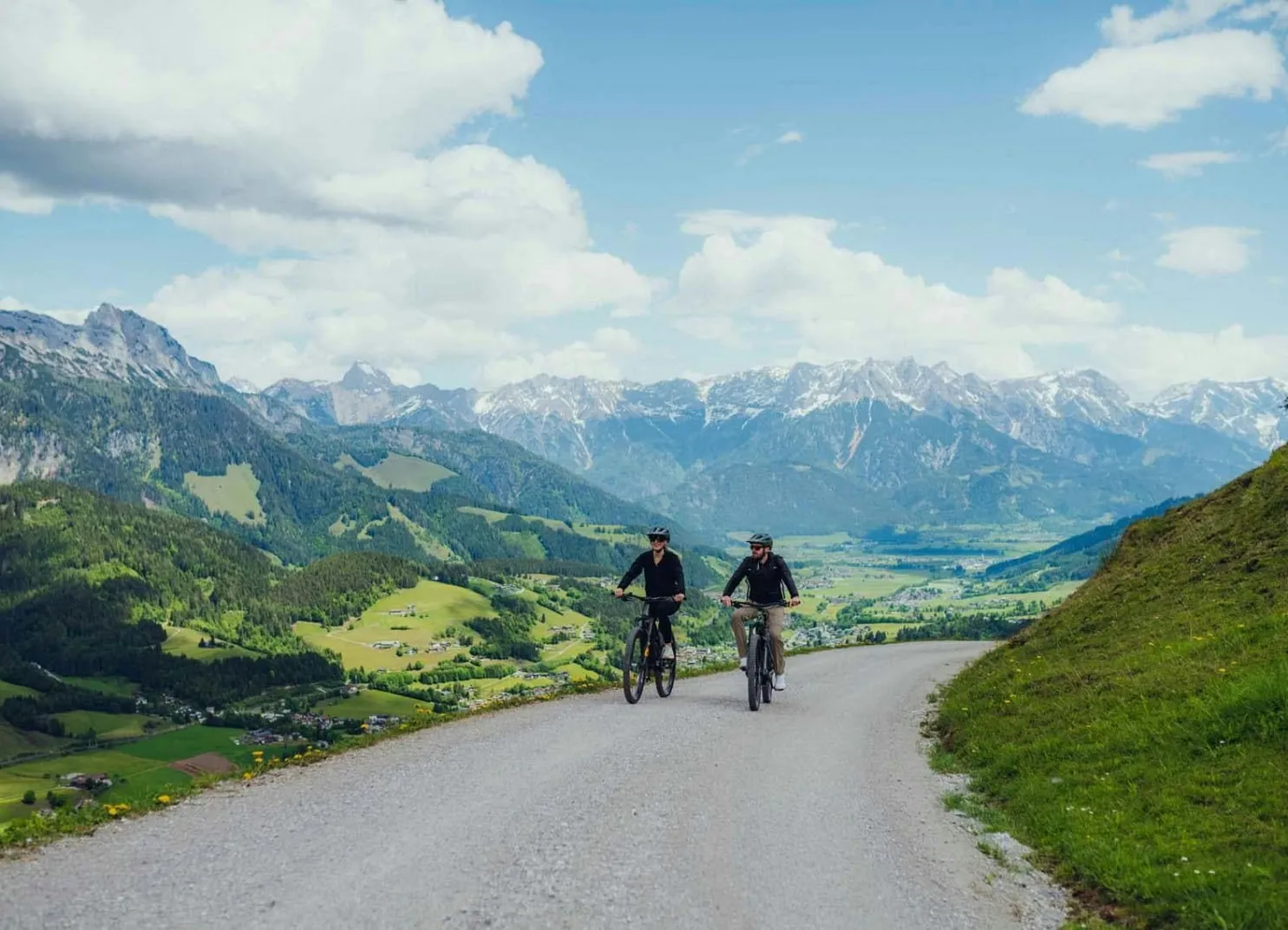 Two cyclists on a road surrounded by mountains and green landscape.